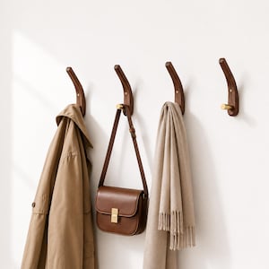 May include: Four wooden wall hooks with brass accents are mounted on a white wall. A tan trench coat, a brown leather purse, and a beige scarf are hanging from the hooks. The hooks have a curved design.