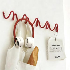 May include: A red metal wall hook with a unique, wavy design. A pair of white and red headphones hangs from the hook, along with a white shirt and a baguette. A small tag with the text "niko and Beache" is also hanging.