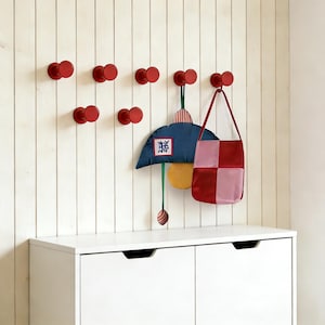 Puede incluir: Ganchos de pared rojos montados en una pared blanca con paneles. Se muestra un adorno de pared con forma de paraguas azul y amarillo con texto japonés, junto con un bolso tote a cuadros rojos y rosas.