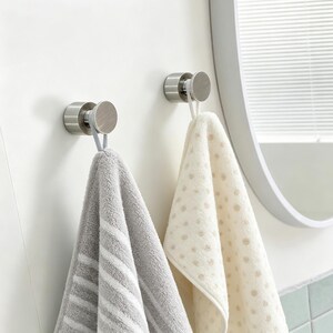 Peut inclure: Deux patères en nickel brossé fixées sur un mur blanc. Chaque patère supporte une serviette : l'une grise à rayures blanches, l'autre crème à petits pois marron. Un miroir rond est visible en arrière-plan. Idéal pour la salle de bain.