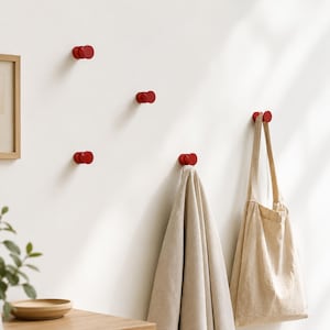 Könnte beinhalten: Mehrere rote Wandhaken sind an einer weißen Wand befestigt. Eine beige Tragetasche und ein beiges Handtuch hängen an zwei der Haken. Die Haken haben ein abgerundetes Doppelkreis-Design. Ein Holztisch und ein gerahmtes Bild sind sichtbar.