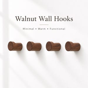 Könnte beinhalten: Vier zylindrische Wandhaken aus dunkelbraunem Walnussholz, die in einer horizontalen Reihe angeordnet sind. Der Text darüber lautet Walnut Wall Hooks, Minimal, Warm, Functional. Jeder Haken hat eine abgerundete Vorderseite und eine glatte Maserung.