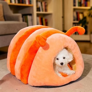 May include: A plush, soft pet bed shaped like a caterpillar with a rounded, tunnel-like entrance. The exterior features alternating bands of light peach and vibrant orange fabric, with two small, curved orange antennae extending from the top.