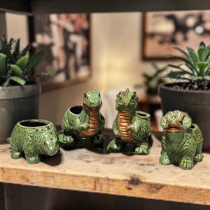 May include: Four green ceramic dinosaur planters with a textured finish. The planters feature various dinosaur designs, including a triceratops, a T-Rex, and a stegosaurus. The planters are displayed on a wooden shelf.