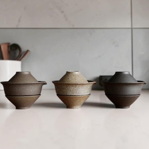 Könnte beinhalten: Drei keramische Gaiwan-Teesets in einer Reihe angeordnet. Jedes Set besteht aus einer Schale mit ausgestelltem Rand und einem passenden konischen Deckel. Das linke Set ist dunkelbraun, das mittlere hellbraun gesprenkelt und das rechte anthrazitfarben.