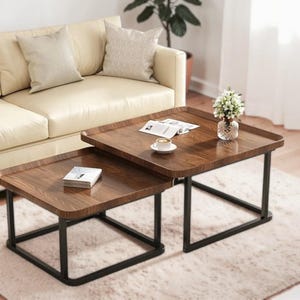 May include: Two nesting coffee tables with dark wood tops and black metal legs. The tables are rectangular with rounded corners. A small vase of flowers and a cup of coffee sit on the larger table. A book and a magazine are also on the tables.