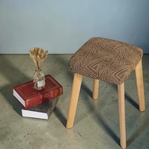 Puede incluir: Un pequeño taburete de madera con un asiento cuadrado con un patrón geométrico marrón. El taburete tiene cuatro patas de madera de color claro. Junto al taburete hay dos libros y un pequeño jarrón de cristal con flores secas.
