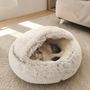 Op de afbeelding: Een zacht, rond, lichtgrijs kattenbed met een kapontwerp. Het bed heeft een pluizige, imitatiebont buitenkant en een gezellig interieur. Een kat ligt opgerold in het bed en slaapt vredig. Het bed staat op een lichtgrijs tapijt.