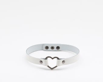 Puede incluir: Una gargantilla blanca con un anillo metálico central en forma de corazón de tono plateado. La banda está asegurada con dos remaches plateados a cada lado del corazón e incluye una correa ajustable con tres botones de presión metálicos.