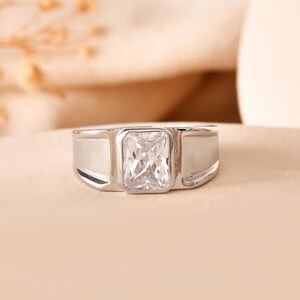 Könnte beinhalten: Ein silberner Ring mit einem rechteckigen, klaren Edelstein in der Mitte. Der Ring hat ein breites Band mit poliertem Finish. Der Edelstein hat eine helle, klare Farbe, und der Ring ist vor einem neutralen Hintergrund platziert.