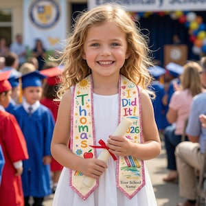 May include: A young girl smiles, wearing a graduation sash that says "I Did It!" and "Thomas" with colorful illustrations. She holds a rolled diploma tied with a red ribbon, celebrating her elementary school graduation.