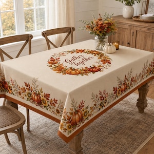 May include: A rectangular beige tablecloth featuring a central wreath of autumn leaves, sunflowers, and pumpkins with the text Gather Together in the middle. The border is decorated with a repeating pattern of orange pumpkins and fall foliage.