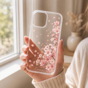May include: Clear phone case with a floral design featuring pink cherry blossoms. The case has the name "Emma" printed on it. The design includes scattered petals and a branch of flowers.