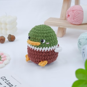 Może przedstawiać: Ręcznie robiona szydełkowa kaczka w odcieniach zieleni, brązu i bieli. Kaczka ma żółty dziób i stopy oraz czarne oczy. Przedmiot rzemieślniczy jest prawdopodobnie elementem dekoracyjnym lub zabawką.