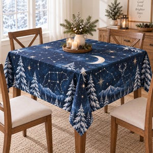 May include: A square tablecloth featuring a dark blue night sky pattern with white constellations, a crescent moon, and a winter forest landscape with snow-covered pine trees, a bear, and a deer silhouette printed across the fabric surface.