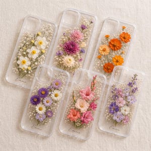 May include: Clear phone cases with pressed flowers in various colors, including white, yellow, orange, pink, purple, and lavender. Each case features a unique floral arrangement with delicate baby's breath. The name "Emma" is printed on the bottom of each case.