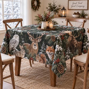 May include: A square tablecloth featuring a dark green forest scene with a snowy owl, a deer, a moose, a fox, a rabbit, and a squirrel. The fabric is decorated with pine branches, pinecones, and red berries. A white pitcher with greenery sits on top.