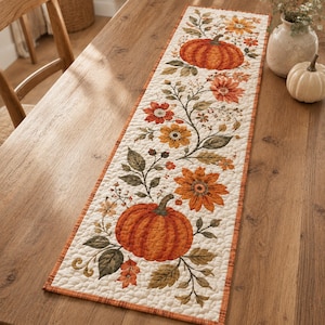 May include: A rectangular quilted table runner featuring a cream-colored background with a printed pattern of orange pumpkins, brown and orange flowers, green leaves, and thin vines. The runner has a thin orange border and a textured stitched finish.