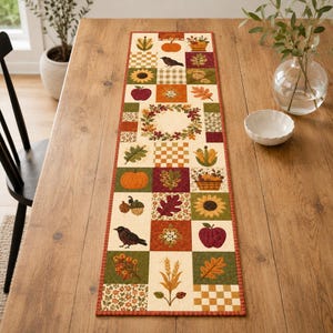 May include: A rectangular fabric table runner featuring a patchwork design of autumn-themed squares. The pattern includes illustrations of pumpkins, sunflowers, apples, acorns, wheat stalks, oak leaves, and black birds in shades of orange, red, and green.