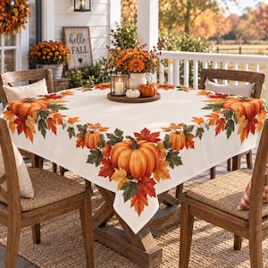 May include: A square off-white tablecloth featuring a printed border of orange pumpkins and multicolored autumn leaves. The design includes large pumpkins at the corners and smaller pumpkins along the edges, set on a wooden table with chairs.