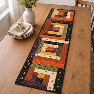 May include: A rectangular quilted table runner featuring a log cabin block pattern in shades of orange, black, green, and cream. The fabric displays small printed motifs of pumpkins, black cats, stars, and crescent moons with a black border.