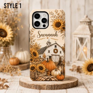May include: A phone case featuring a rustic farm scene with a white barn, sunflowers, pumpkins, and a lantern. The name Savannah is printed in a script font above the barn. The design includes autumn foliage and a plaid fabric texture at the base.