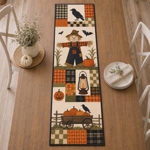 May include: A rectangular table runner featuring a patchwork design with autumn-themed illustrations including a scarecrow, a lantern, pumpkins, a wooden wagon, and black crows, accented by orange, black, and green plaid and checkered patterns.