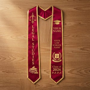 May include: Burgundy graduation sash with gold trim. The sash features the words "LEGAL STUDIES" and "Class of 2026". Gold graphics include a gavel, scales of justice, books, and a graduation cap. The sash is designed to be personalized with a name and law school.