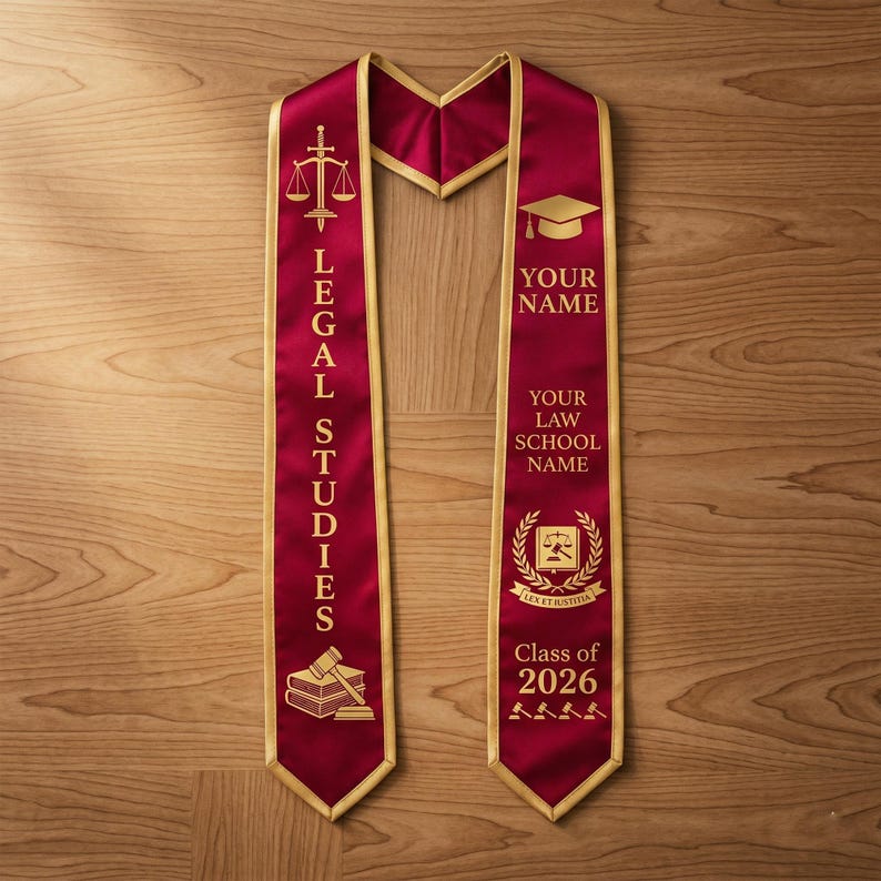 May include: Burgundy graduation sash with gold trim. The sash features the words "LEGAL STUDIES" and "Class of 2026". Gold graphics include a gavel, scales of justice, books, and a graduation cap. The sash is designed to be personalized with a name and law school.