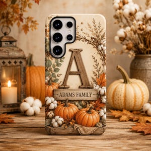 May include: A phone case featuring a large wooden letter A above a rectangular sign with the text ADAMS FAMILY. The design includes orange pumpkins, white cotton bolls, and autumn leaves on a cream background, surrounding the camera lens area.