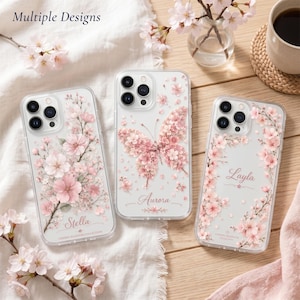 May include: Three clear smartphone cases featuring cherry blossom designs. The left case shows a branch with pink flowers and the name Stella. The center case displays a butterfly made of blossoms and the name Aurora. The right case has a floral border and the name Layla.