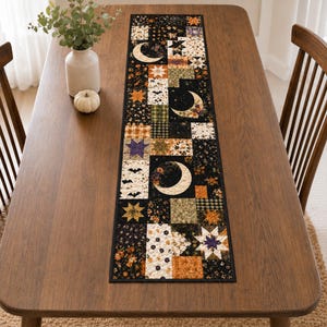 May include: A rectangular quilted table runner featuring a patchwork design of black, orange, cream, and green fabrics. The pattern includes motifs of crescent moons, stars, bats, and floral prints, finished with a solid black fabric border.