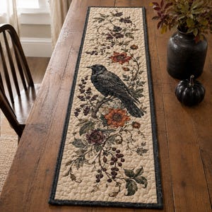 May include: A rectangular quilted table runner featuring a cream-colored background with a central black crow perched on a branch. The design includes detailed illustrations of dark red berries, green leaves, and orange flowers with a dark border.