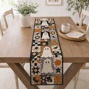 May include: A rectangular quilted table runner featuring a patchwork design with repeating motifs of white ghosts, orange pumpkins, black stars, and bats. The fabric blocks are arranged in a vertical pattern with a black border trim.