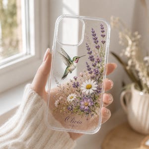 May include: Clear phone case featuring a hummingbird in flight, surrounded by lavender, daisies, and other purple and white flowers. The case has the name "Olivia" printed in script at the bottom. The design is set against a clear, glittery background.