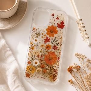 May include: Clear phone case with embedded dried flowers in shades of orange, yellow, and white. The case features a variety of floral elements, including daisies and maple leaves. The name "Emma" is written in script at the bottom. The case is on a white surface.