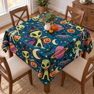May include: A square tablecloth featuring a dark teal background with a repeating pattern of green aliens dressed as witches, mummies, and skeletons. The design includes orange jack-o'-lanterns, yellow crescent moons, stars, bats, planets, and UFOs.
