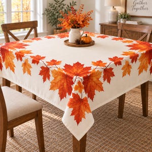 May include: A square tablecloth featuring a cream-colored base with a border of vibrant orange, red, and yellow maple leaves. The design includes a central white pitcher filled with autumn foliage and a small orange pumpkin on a wooden tray.