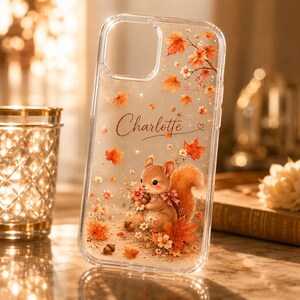 May include: A clear phone case featuring a printed design of a small brown squirrel holding a bouquet of pink flowers, surrounded by scattered orange and red autumn leaves, small acorns, and the name Charlotte written in elegant cursive script.