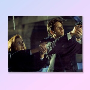 May include: A rectangular print featuring two people pointing handguns. The person on the left has red hair and is wearing a dark suit. The person on the right has dark hair and is also wearing a dark suit. The background is a dimly lit interior.