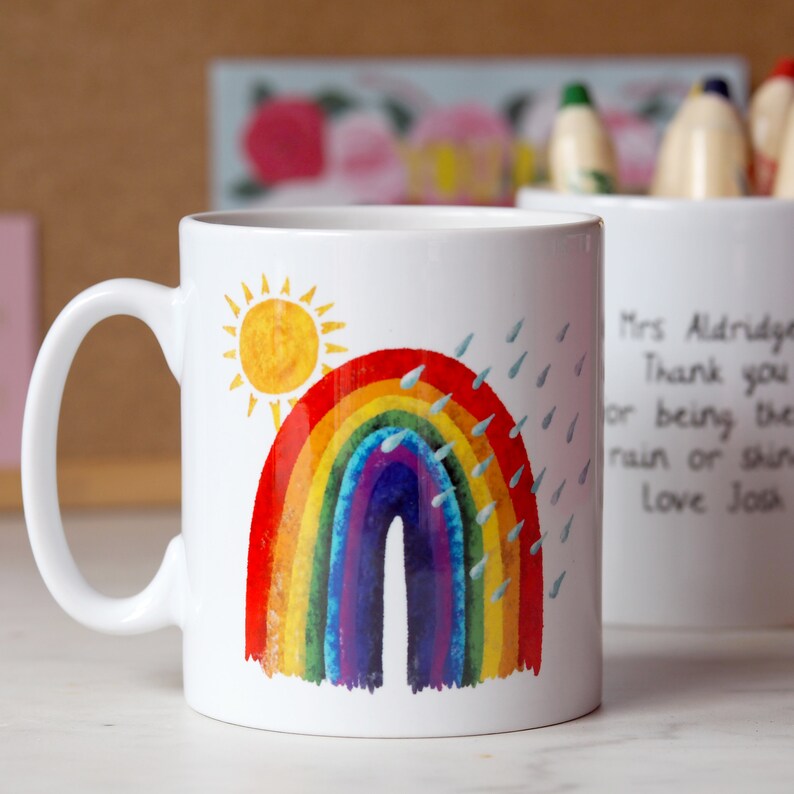 A colourful mug for teacher, with a bright rainbow, sun and rain design created in a naive print style, like a kids book such as the hungry caterpillar. A personal message is on the back The mug is white ceramic.