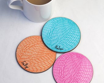 Könnte beinhalten: Drei runde Untersetzer mit einem Garnknäuel-Design in Orange, Blau und Pink, jeweils mit einem Namen: Sally, Helen und Lauren. Im Hintergrund steht eine weiße Tasse mit brauner Flüssigkeit. Die Untersetzer liegen auf einer weißen Oberfläche.