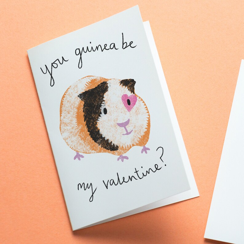 May include: A white greeting card featuring a printed illustration of a brown, black, and white guinea pig with a pink heart shape over one eye. The card includes the black handwritten-style text "you guinea be my valentine?" printed on the front.