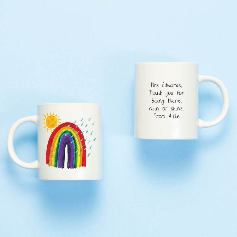 May include: Two white ceramic mugs. One mug has a rainbow, sun, and raindrops drawn on it. The other mug has the text "Mrs Edwards. Thank you for being there. rain or shine. From Alfie" written on it.