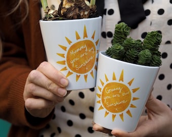 May include: Two white ceramic flower pots with yellow sun designs and handwritten messages. The first pot says "Mummy you are my Sunshine" and the second pot says "Granny you are our Sunshine".
