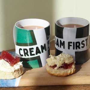May include: Two ceramic mugs with tea, featuring the words "CREAM" and "JAM FIRST". The mugs have a white base with green and black geometric patterns. Scones with cream and jam are placed on a wooden board, beside the mugs.