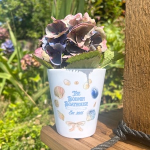 May include: A white ceramic flower pot decorated with seashells and the text "The Bodmin Boathouse Est. 2025". The pot holds a bouquet of purple and pink hydrangeas. The pot is placed on a wooden surface.