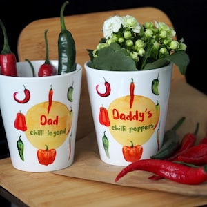 May include: Two white ceramic plant pots with red and green chilli pepper designs. One pot says "Dad chilli legend" and the other says "Daddy's chilli peppers".