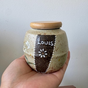 Peut inclure: Petit pot en céramique mouchetée avec un couvercle en bois. Le pot présente un patch marron foncé avec le nom "Louis" et un motif floral blanc. Le pot est tenu dans une main.