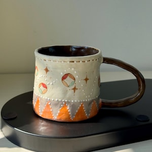 Puede incluir: Una taza de cerámica hecha a mano con asa e interior marrón. El exterior es blanquecino con un patrón de círculos naranjas, rojos y verdes, estrellas marrones y puntos blancos. La base presenta triángulos naranjas y grises.